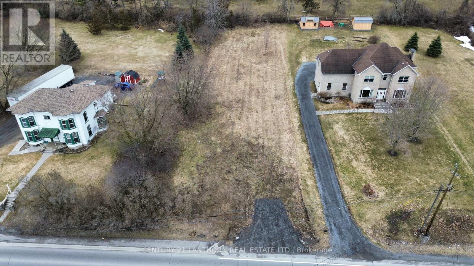 Aerial view of a vacant residential lot located between two existing homes, with road frontage, open grassy space, mature trees, and a private driveway beside a neighbouring estate-style home.